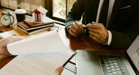 A man in a suit is sitting at a desk with a laptop and a model house.
