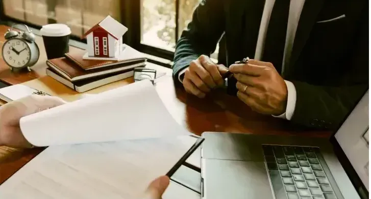 A man in a suit is sitting at a desk with a laptop and a model house.