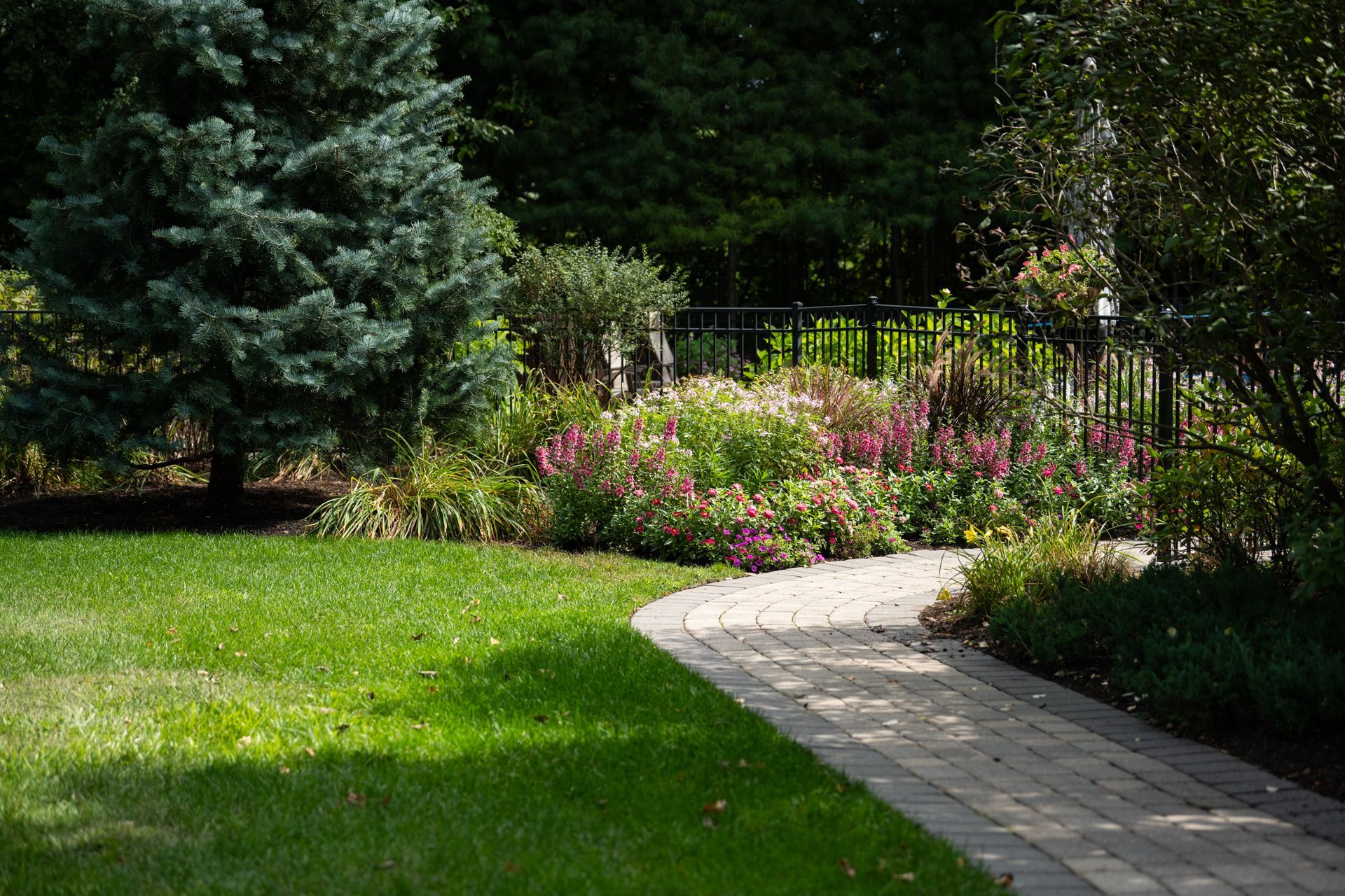 A brick walkway leading to a lush green yard surrounded by trees and flowers.