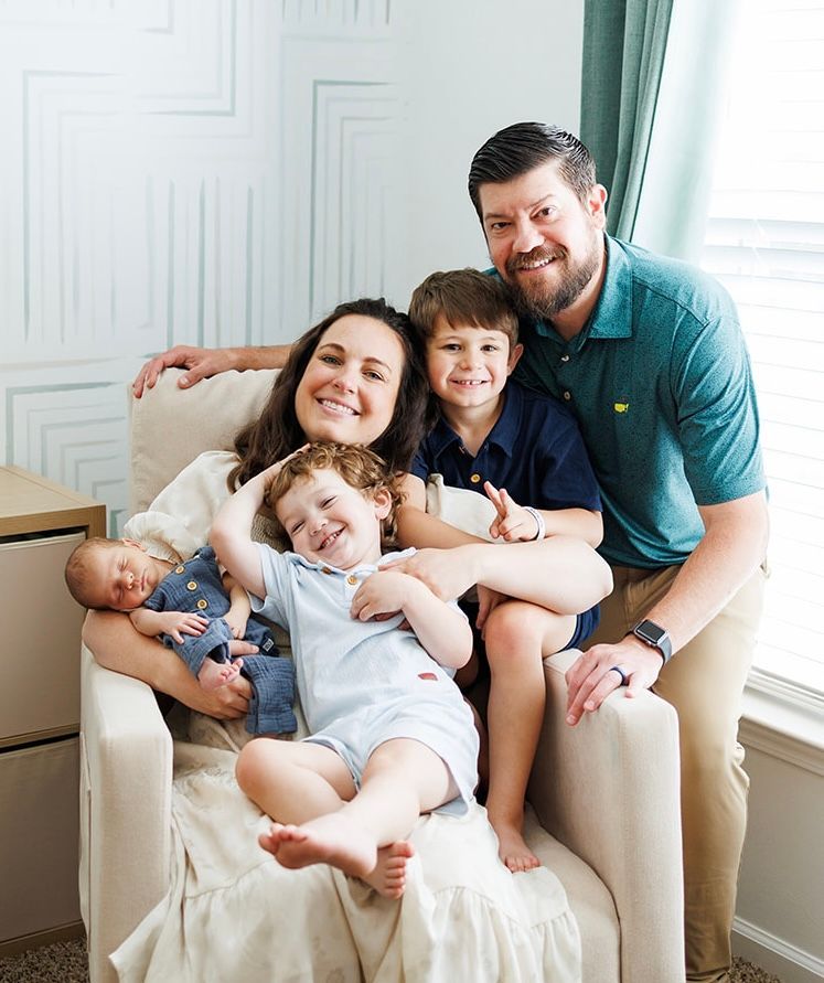 Family of four sitting together on a cream armchair, smiling in a bright room by a window.