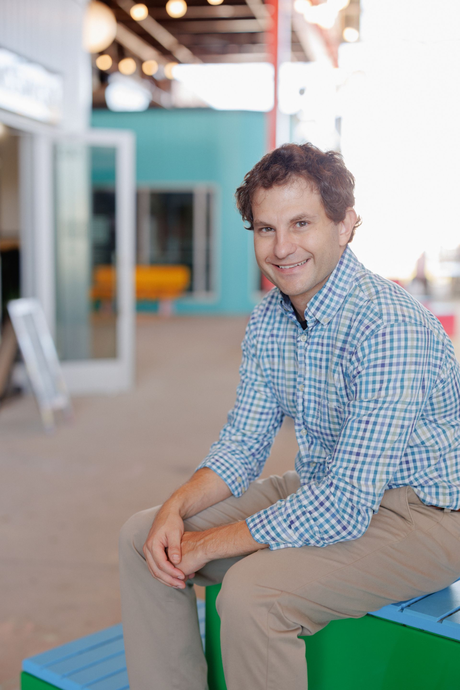 Smiling man in a blue checkered shirt sitting on a green bench in a bright indoor space