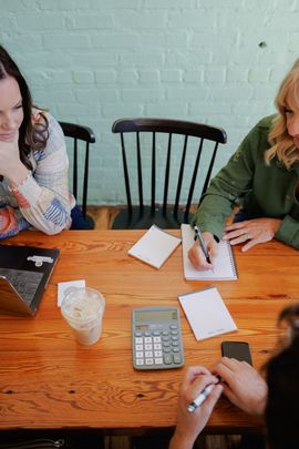 People writing on notepads at a wooden table with a calculator and drinks