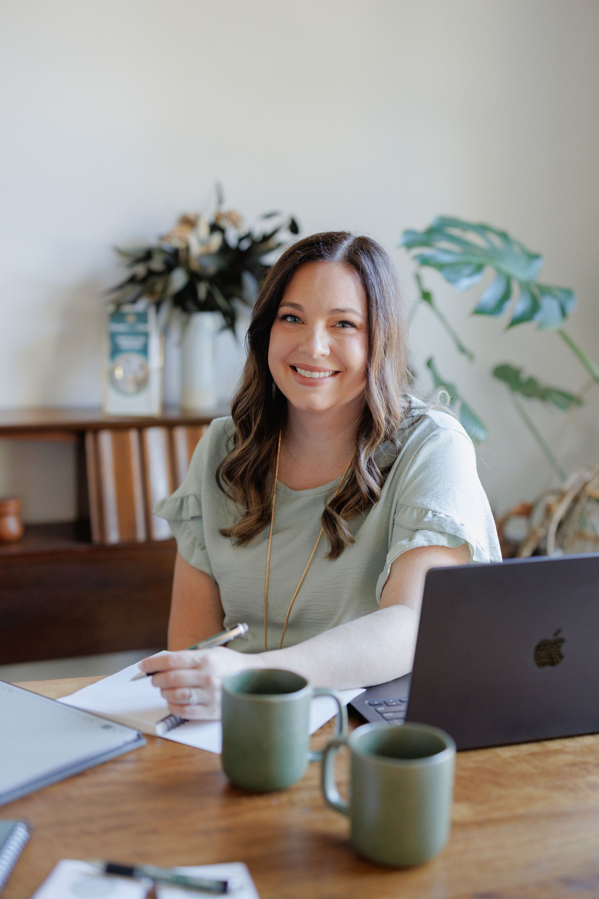 Woman smiling at a desk with a laptop, mug, and notebook in a bright home office