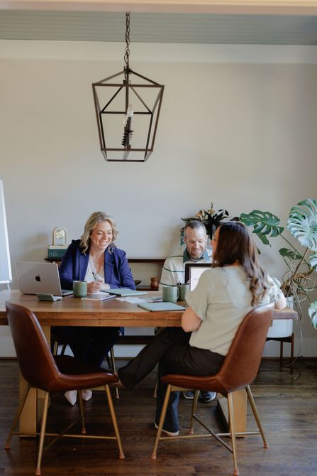 Three people talking at a wooden dining table in a bright room with hanging geometric light fixture.
