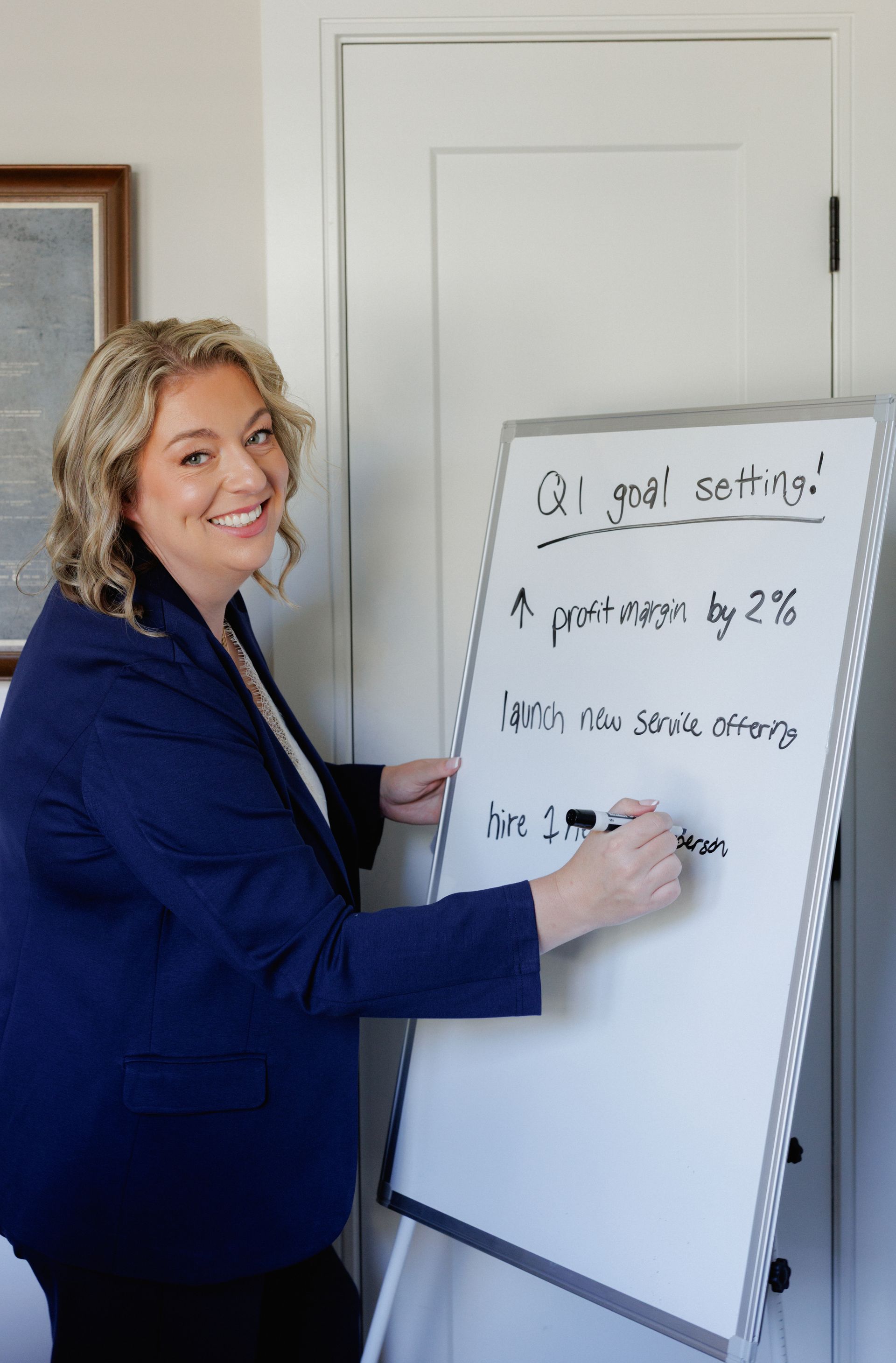 Woman in blue blazer pointing at a whiteboard with handwritten notes in an office.