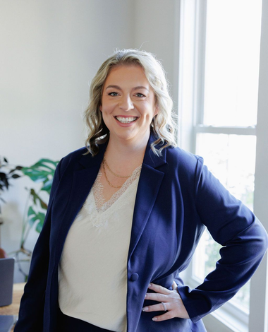 Smiling woman in a navy blazer and white blouse standing with hand on hip in a bright office.