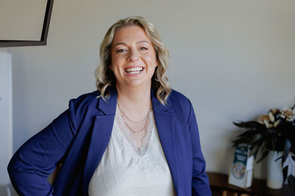 Smiling person in a blue blazer standing indoors beside a plant.