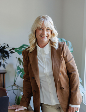 Smiling person in a brown blazer and white blouse standing indoors near a plant.