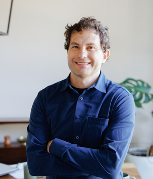 Smiling person in a blue shirt with arms crossed, standing indoors near a plant and shelf.