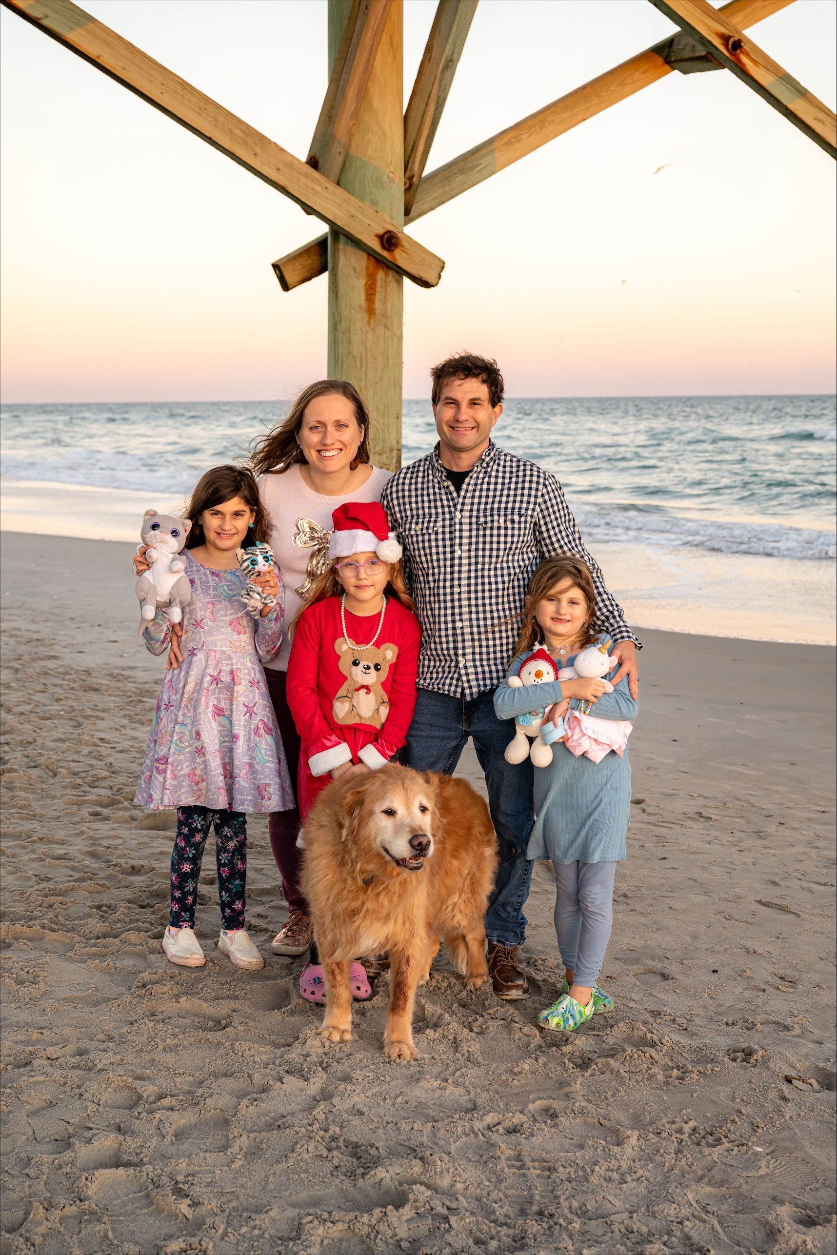 Family standing on a beach at sunset with a golden retriever under wooden pier beams