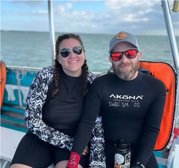 Two people smiling on a boat, wearing sunglasses with the ocean behind them.