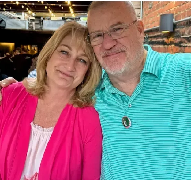 Two smiling people posing together indoors, wearing pink and teal tops.