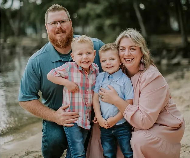 Family of four smiling outdoors by a dirt path, with parents standing behind two young children.