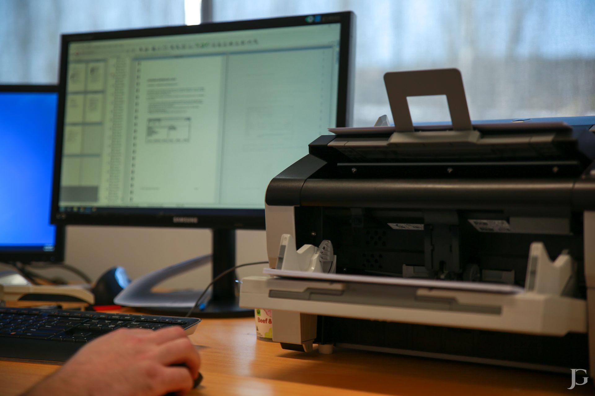 Printer on a desk in front of a computer screen. A hand uses a mouse.