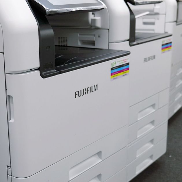 White Fujifilm commercial printer with black accents, stacked paper trays, and color indicator labels.
