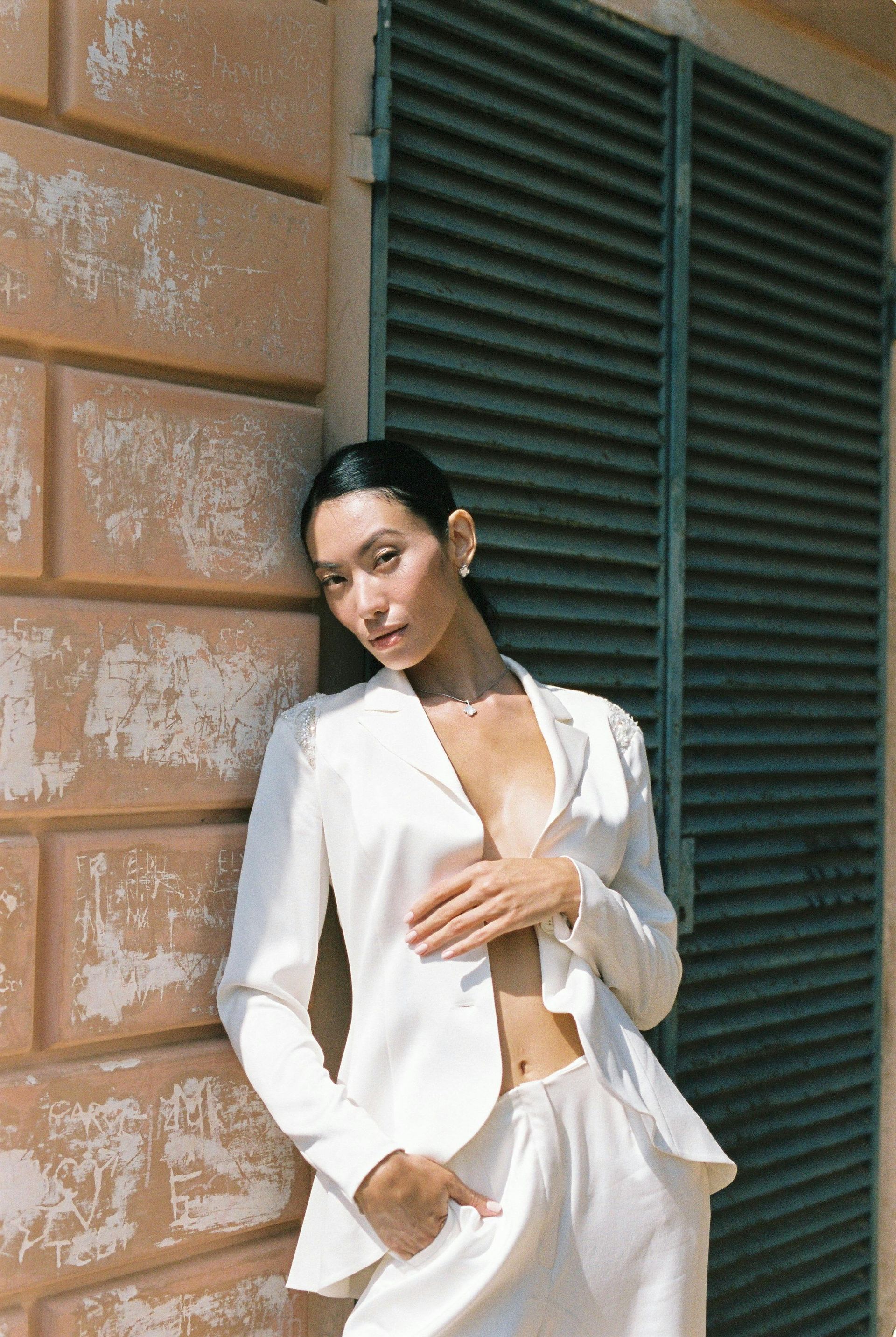 Woman in white suit leans against a brick wall. Sunlight, green shutter backdrop, hand on chest, looking at camera.