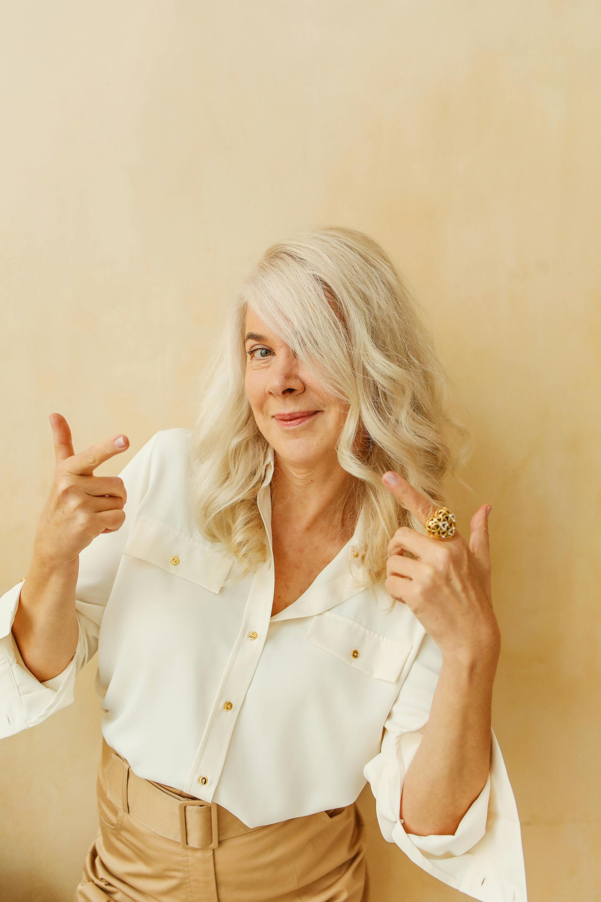 Woman with white wavy hair, pointing to herself with both hands, wearing a white shirt and gold pants.