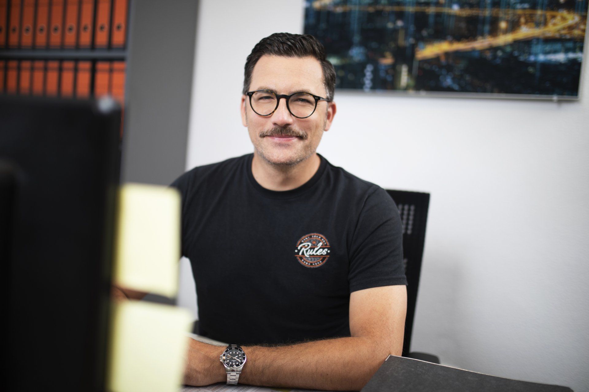 Ein Mann mit Brille und Schnurrbart sitzt an einem Schreibtisch vor einem Computer.