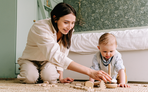 Woman and baby playing with wooden toys on a floor beside a bed