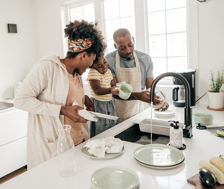 Family washing dishes together in a bright kitchen, with one person loading the sink and another drying plates