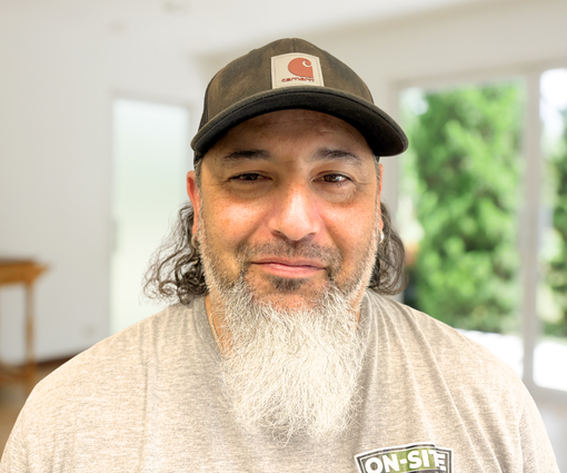 Man wearing a black cap and gray shirt, smiling indoors with a bright blurred background