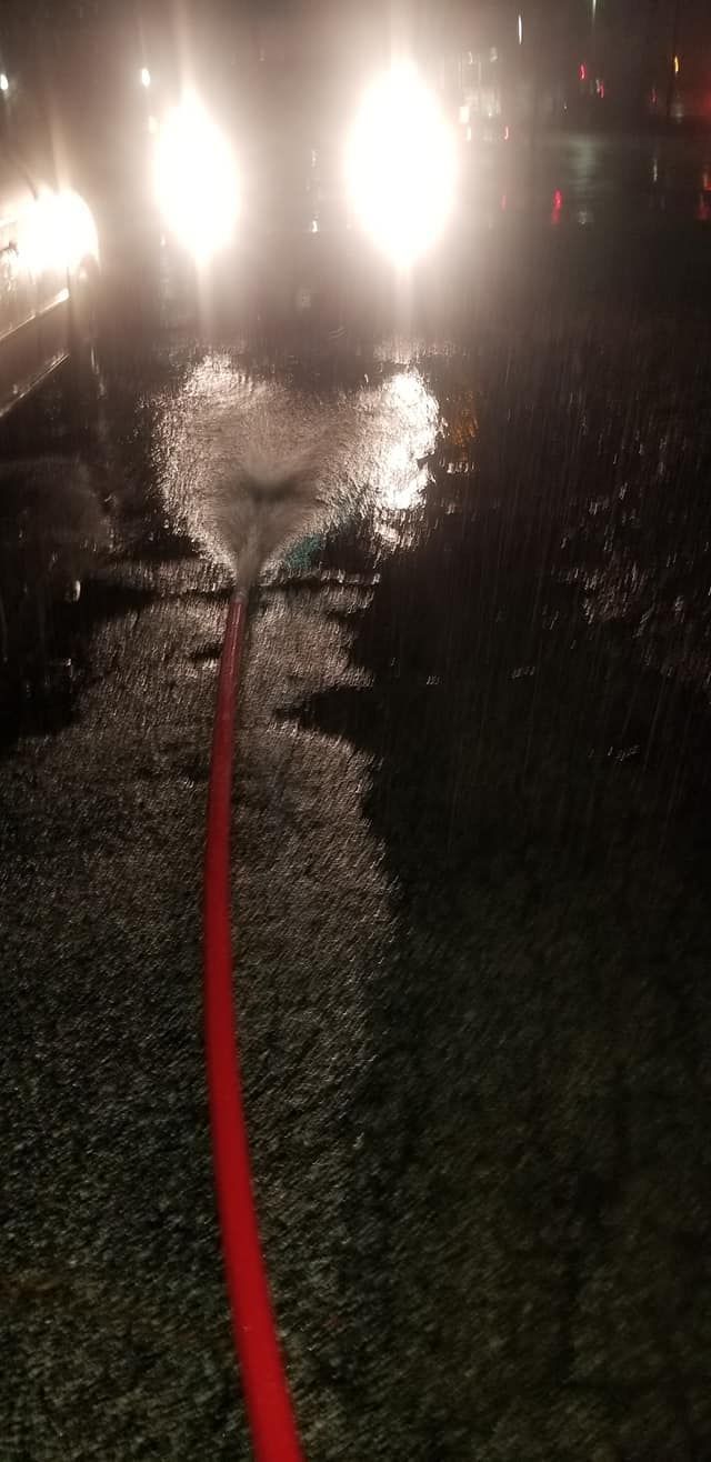 A red hose sprays water on a wet street at night, with headlights in the background.
