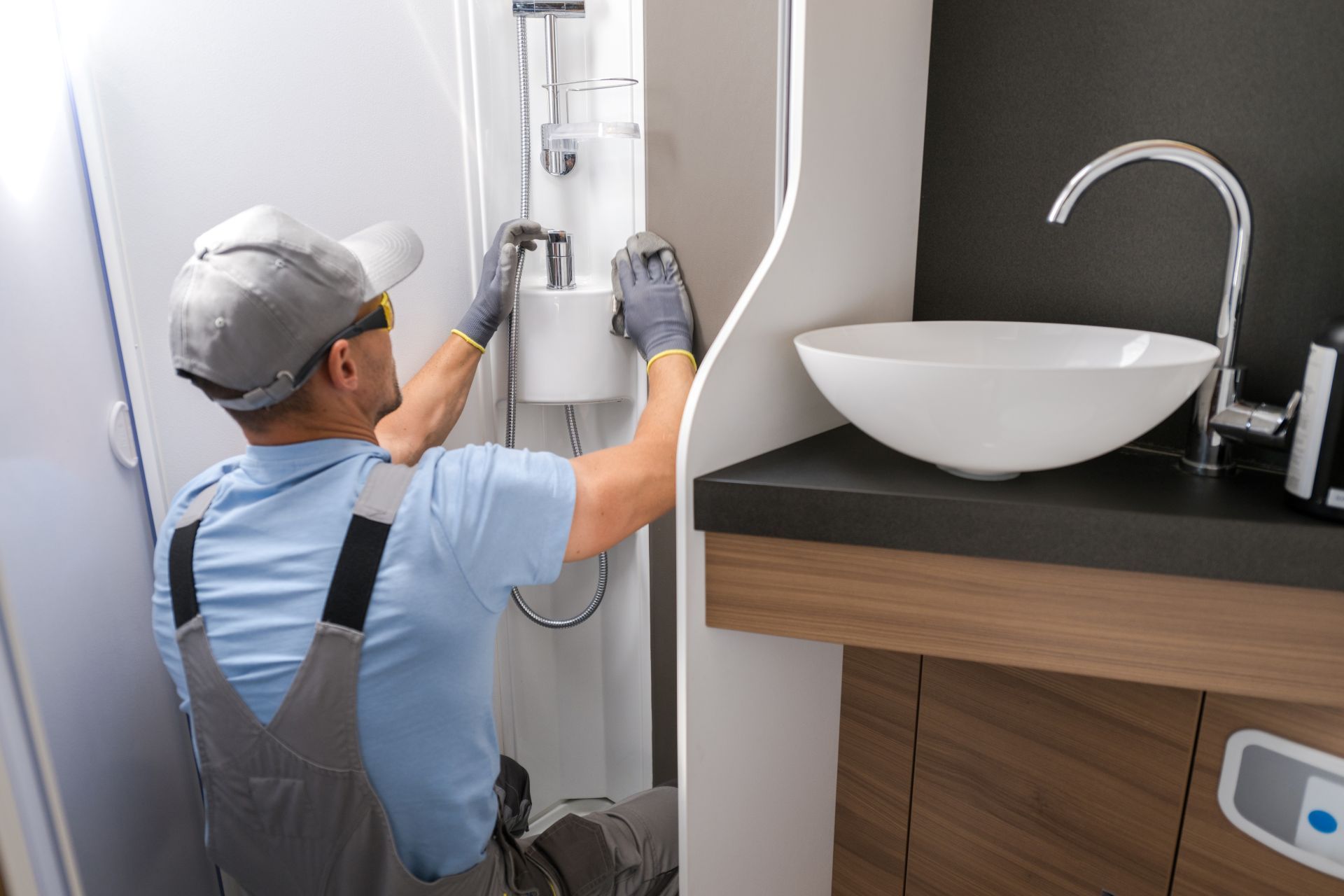 Person in overalls installs shower fixture near a sink in a small RV bathroom.