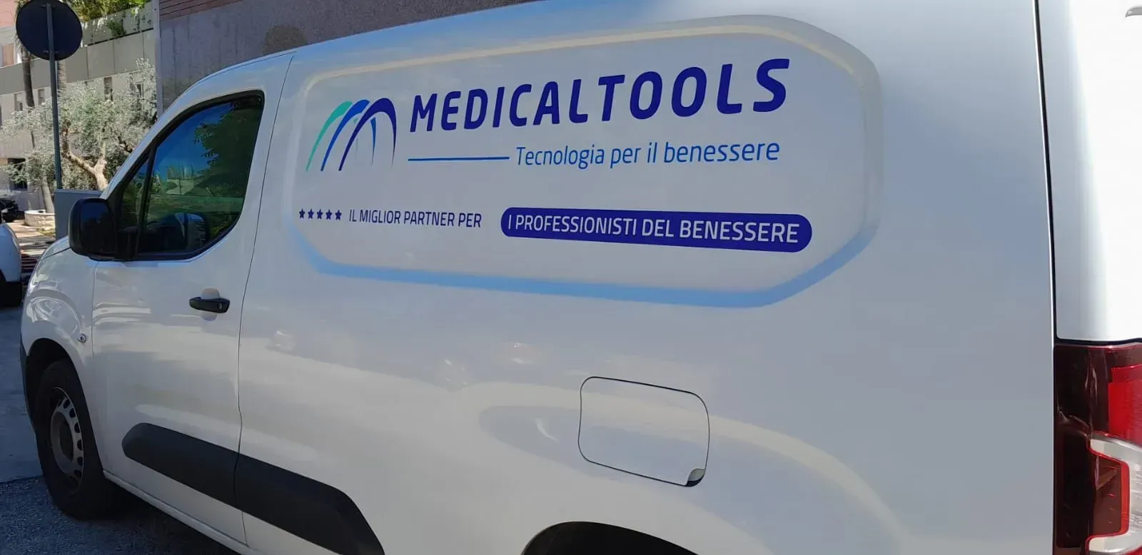 Furgone bianco per la consegna di strumenti medici con logo e testo blu, parcheggiato all'aperto sotto il sole.