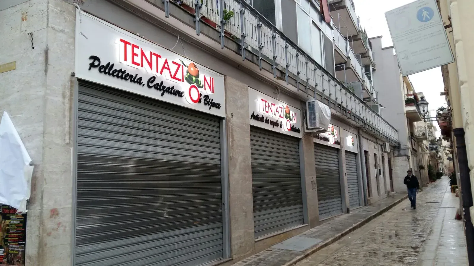 Vetrine chiuse con saracinesche metalliche lungo una stretta strada bagnata in una giornata grigia.