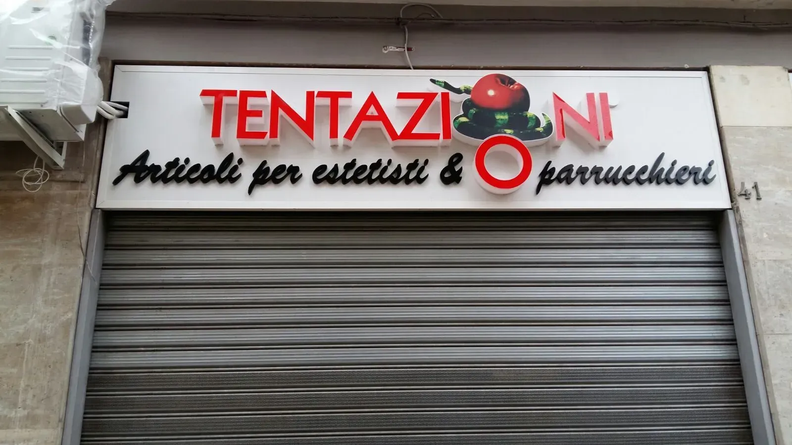 Storefront sign reading “TENTAZIONI” above a closed metal shutter; “Articoli per estetisti & parrucchieri” below.