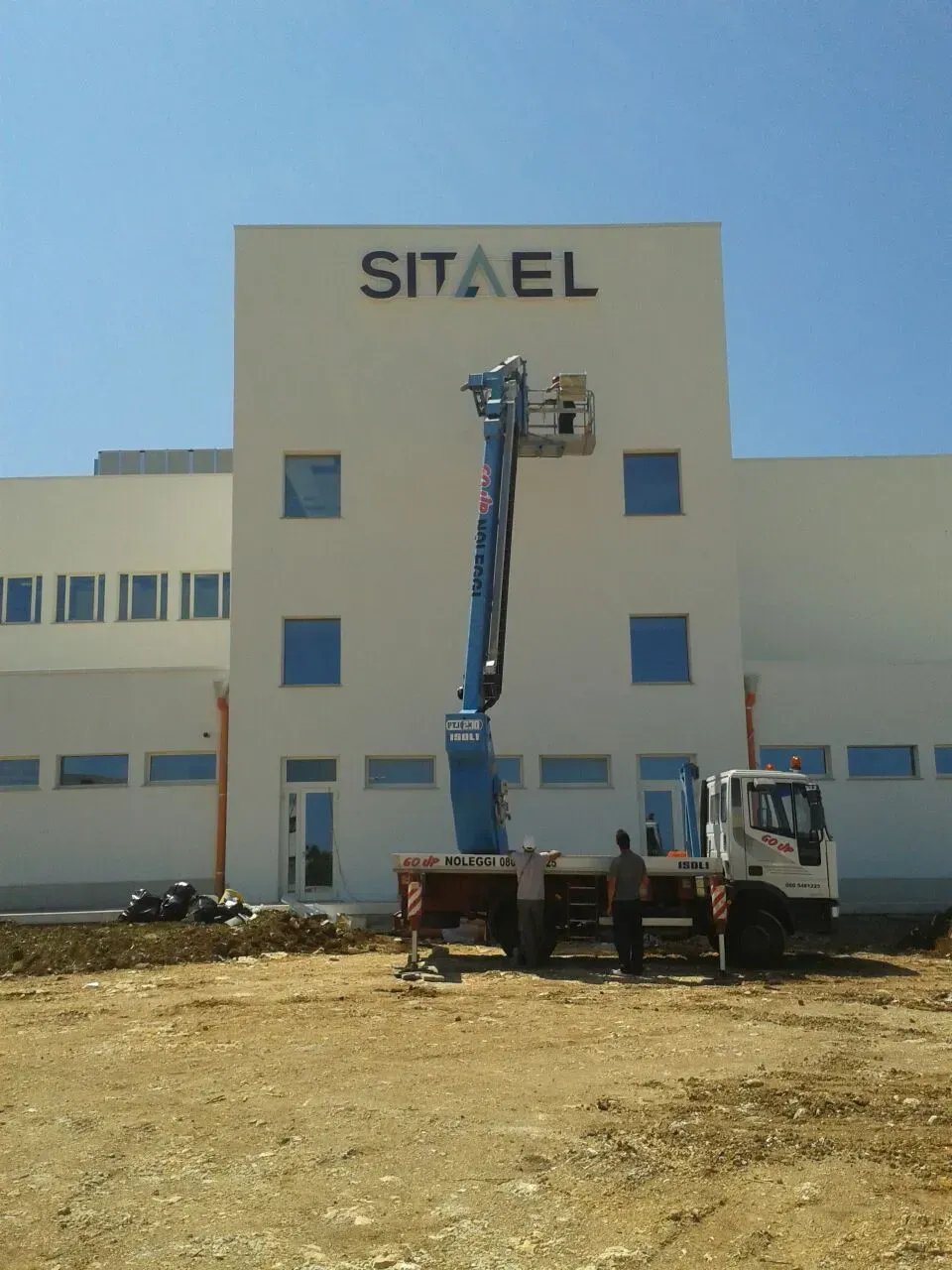 Facciata dell'edificio con l'insegna SITEL e una piattaforma aerea montata su camion in funzione vicino alla finestra più alta.