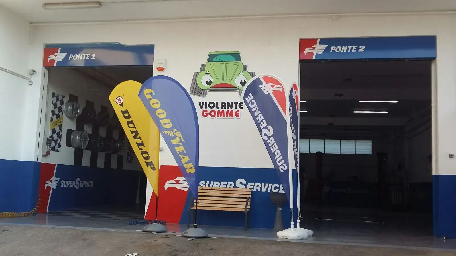 Ingresso di una piccola officina meccanica con striscioni e bandiere blu e rosse e postazioni di lavoro aperte.