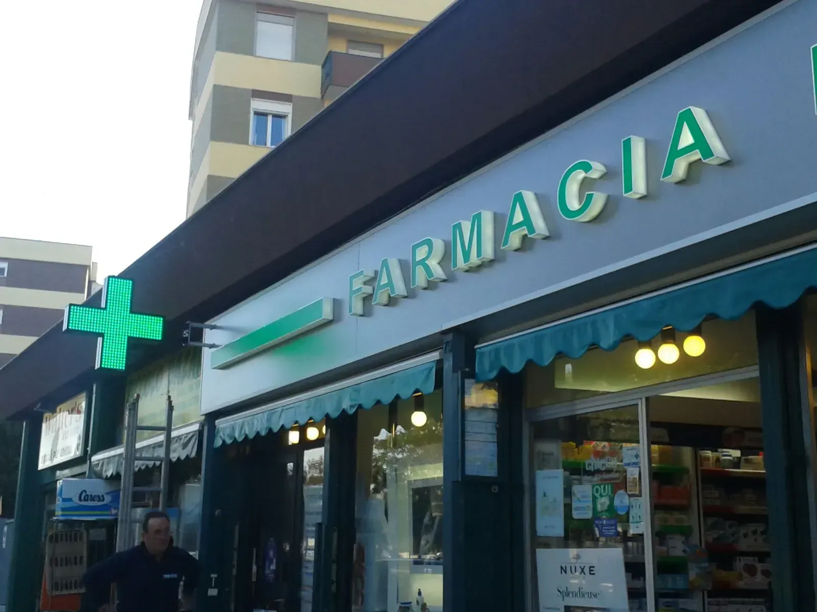 Esterno della facciata di una farmacia con insegna a croce verde e tenda da sole, vista dalla strada.