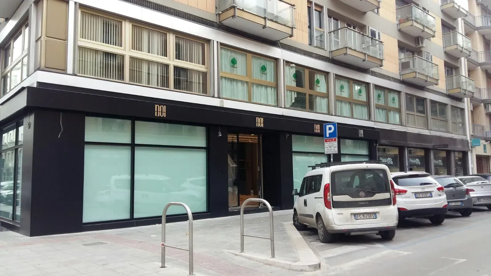 Vista a livello stradale di una vetrina con facciata nera, auto parcheggiate e balconi di appartamenti sovrastanti.