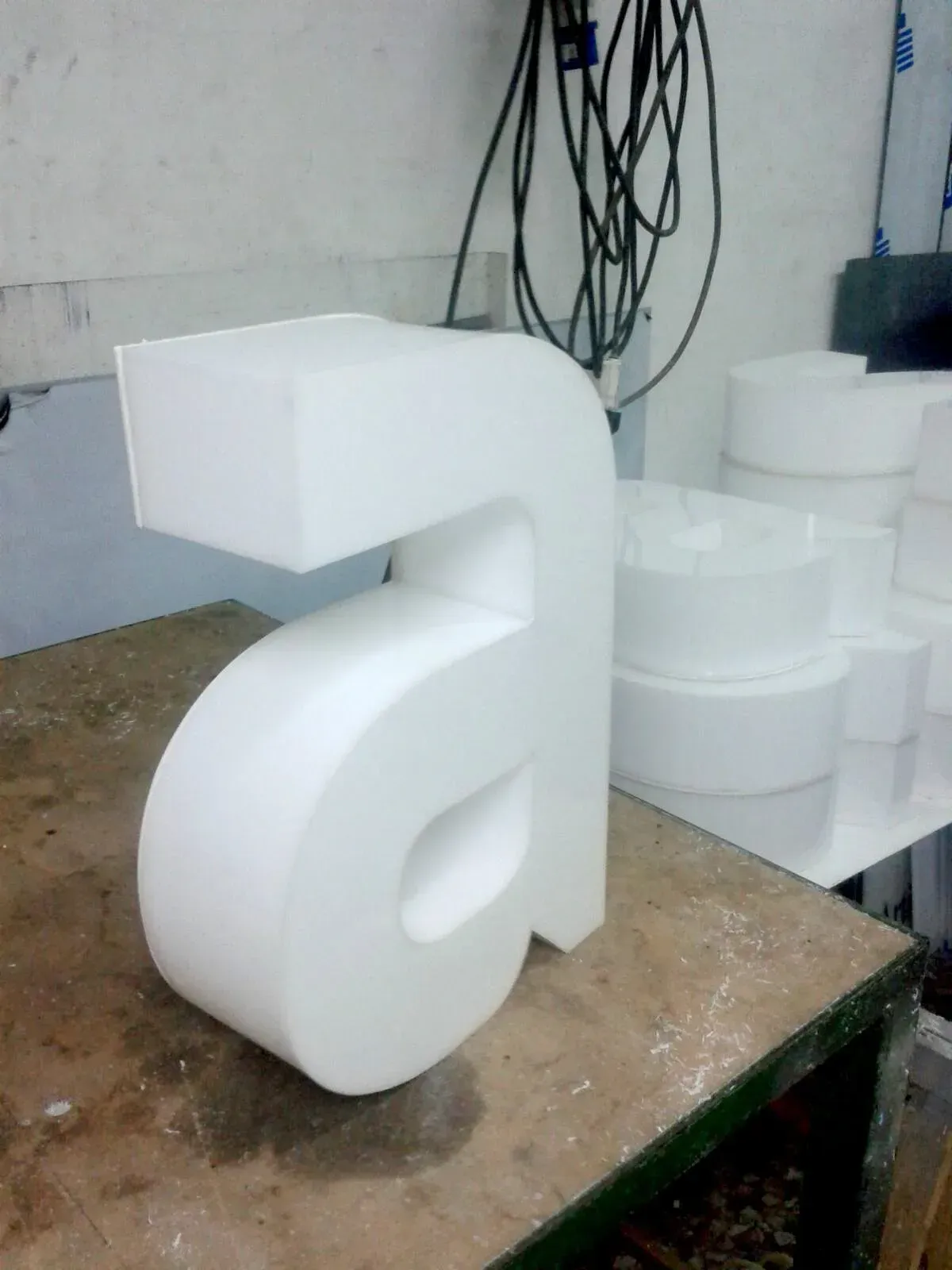 Scultura tridimensionale di lettere in schiuma bianca su un tavolo da lavoro in un'officina, con altre forme in schiuma impilate dietro di essa.