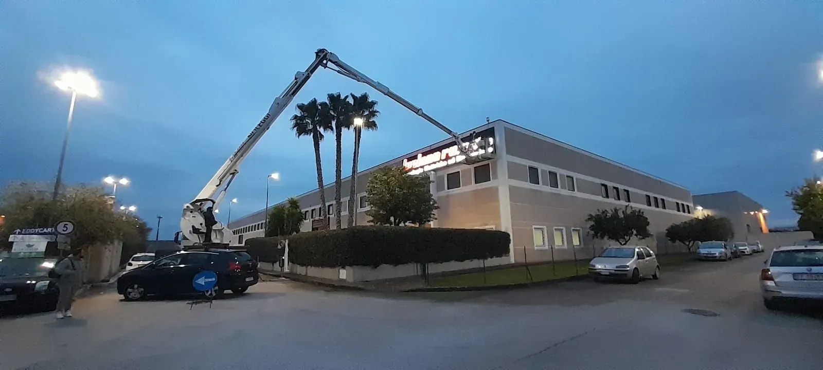 Al crepuscolo, una gru solleva un'insegna luminosa su un edificio commerciale beige, con auto parcheggiate nelle vicinanze.