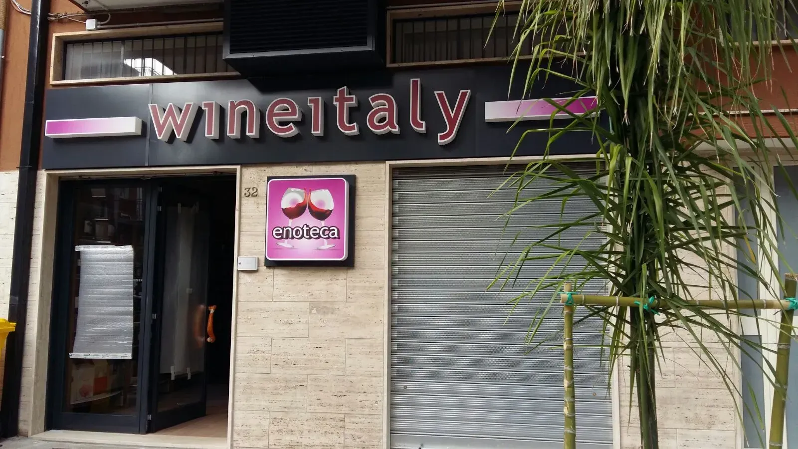 Vetrina del negozio Wineitaly con insegna rosa, ingresso aperto e saracinesca metallica chiusa, accanto a una palma.