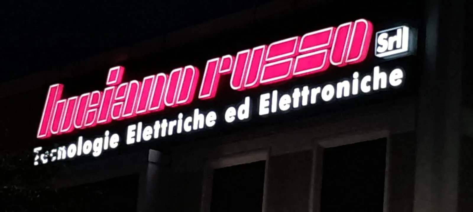 Illuminated pink “Radio Messoa Srl” sign with “Tecnologie Elettriche ed Elettroniche” on a dark building facade