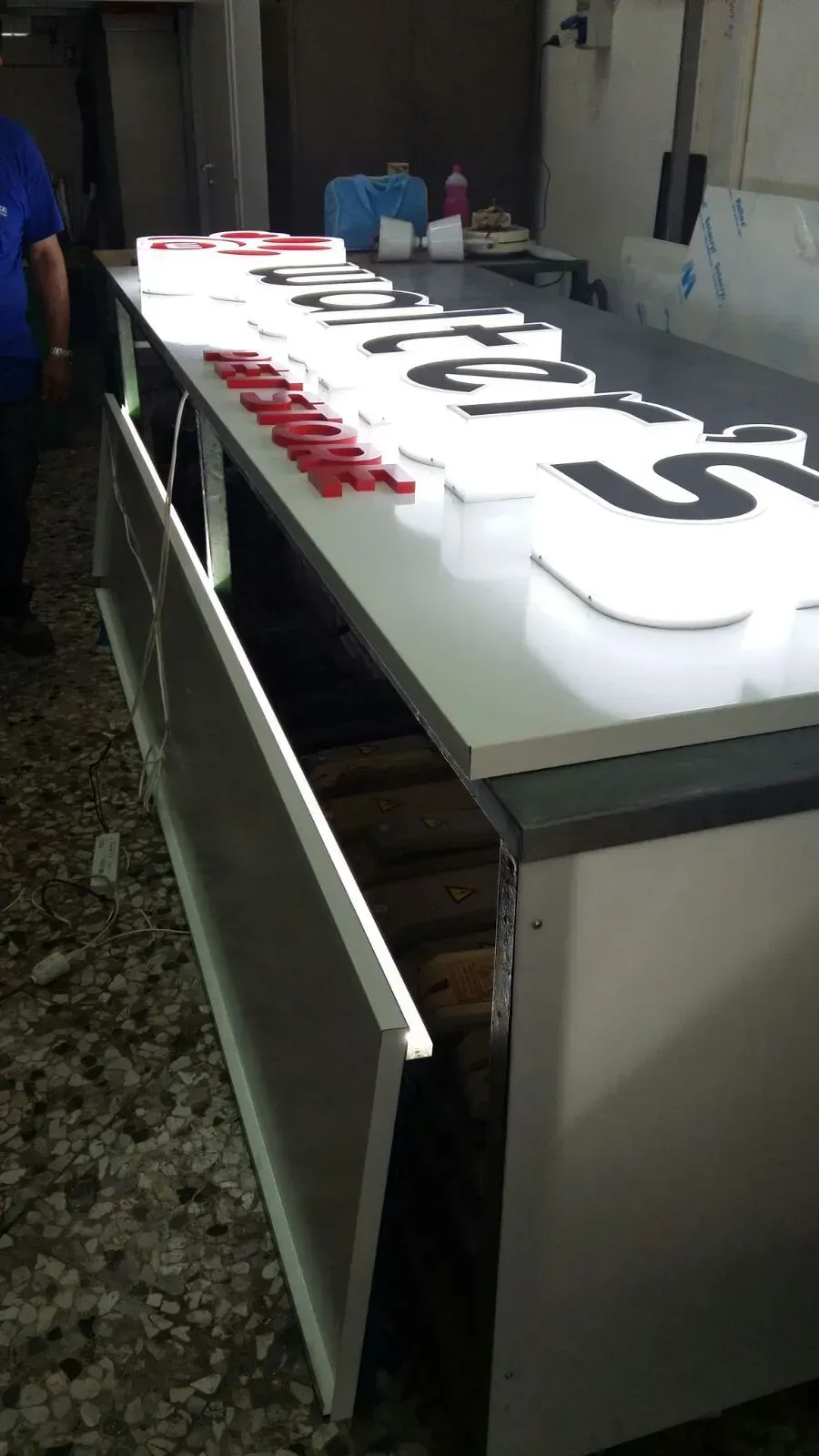 Grande insegna luminosa con scritte nere e rosse su un tavolo, vista di lato in un'officina.
