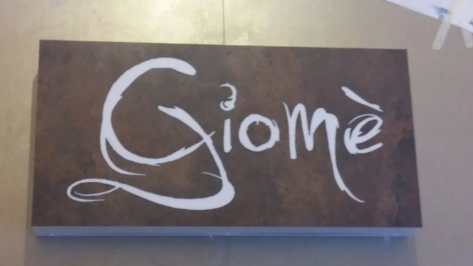 Scritta bianca “Giomé” su un cartello rettangolare marrone, su uno sfondo beige.