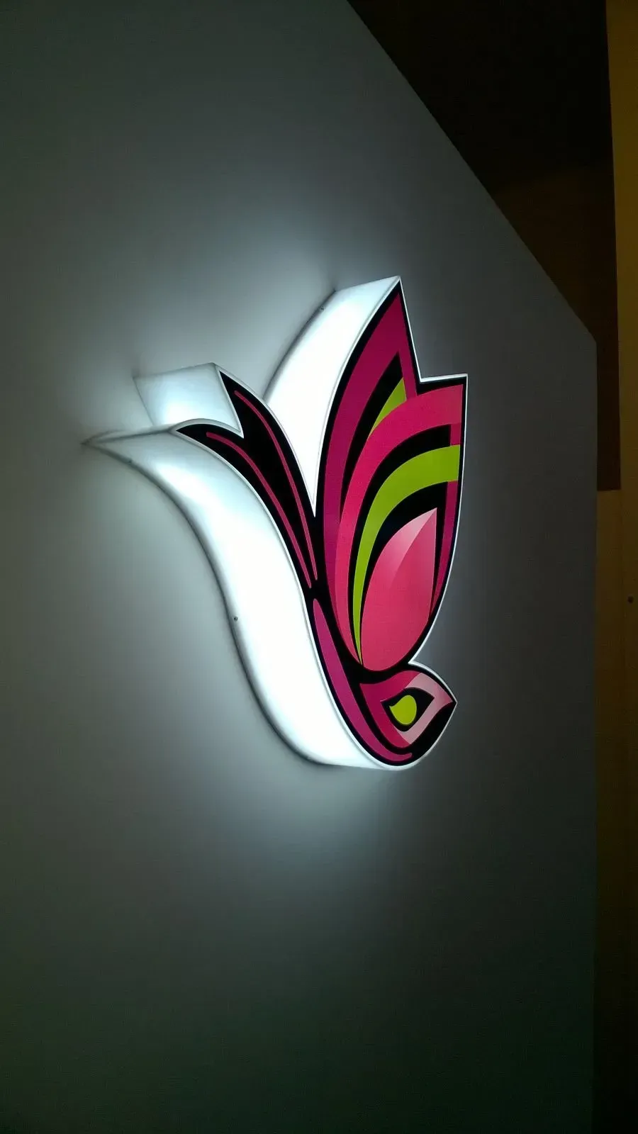 Logo decorativo illuminato rosa, bianco e verde, montato a parete su uno sfondo scuro.