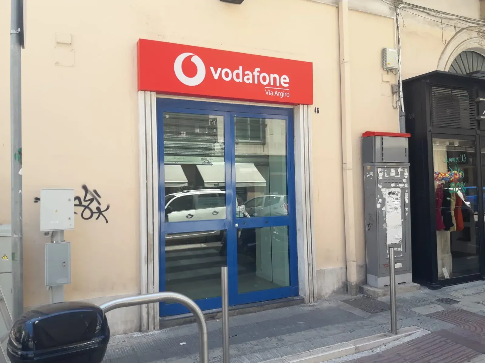 Negozio Vodafone con insegna rossa e porta a vetri blu su una via cittadina.