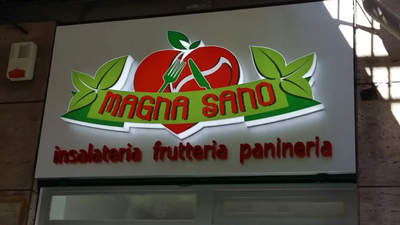 Insegna per la panetteria e pasticceria “MAGNA SANO” con logo a forma di mela rossa e foglie verdi.