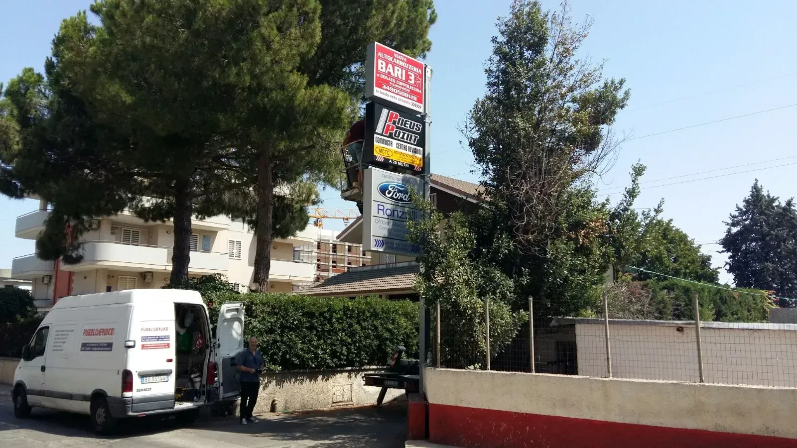 Veduta stradale con un furgone bianco parcheggiato vicino a una siepe e un'insegna alta vicino ad alberi ed edifici.