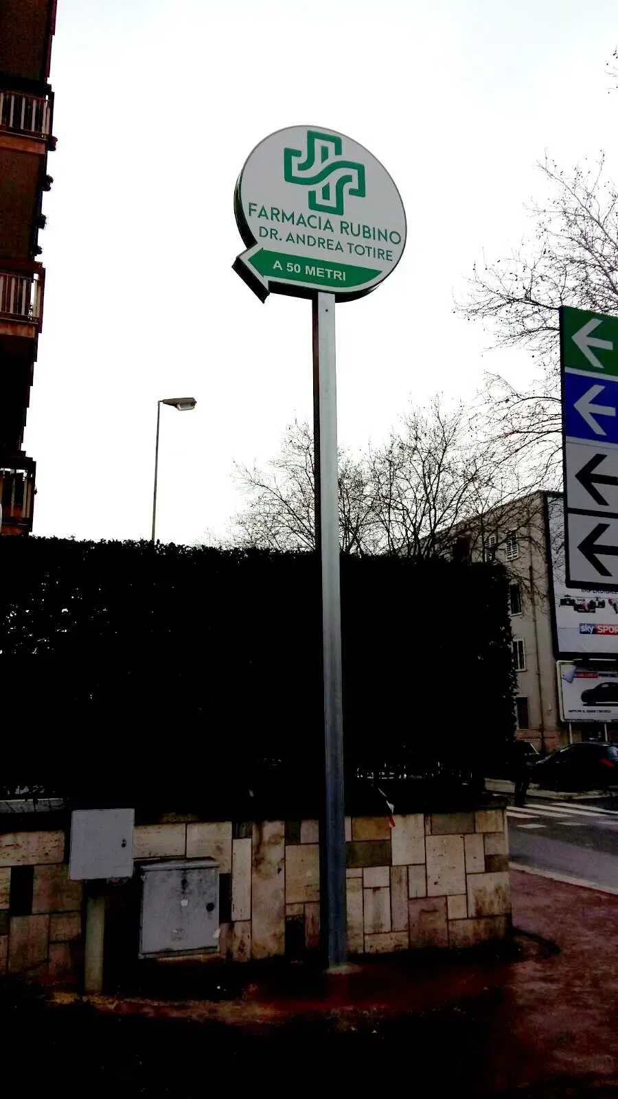 Segnale di fermata dell'autobus all'aperto, su un palo alto, con logo circolare verde e frecce direzionali sulla destra, vicino a un marciapiede.