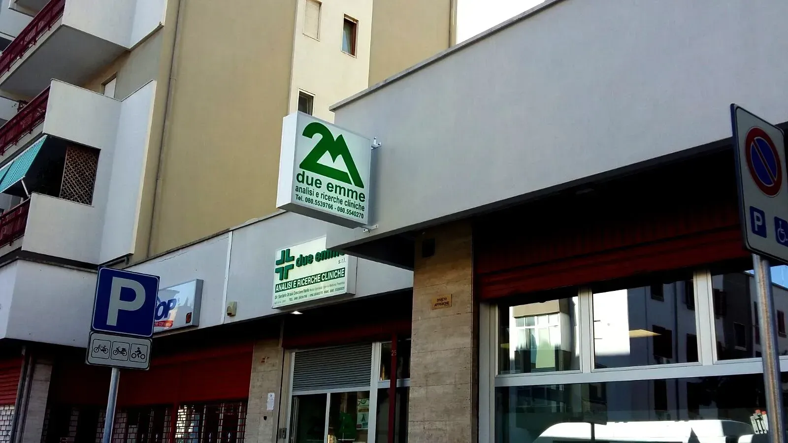 Vetrine che si affacciano sulla strada, complete di insegne, tra cui un'insegna verde per una farmacia e un cartello blu per il parcheggio.