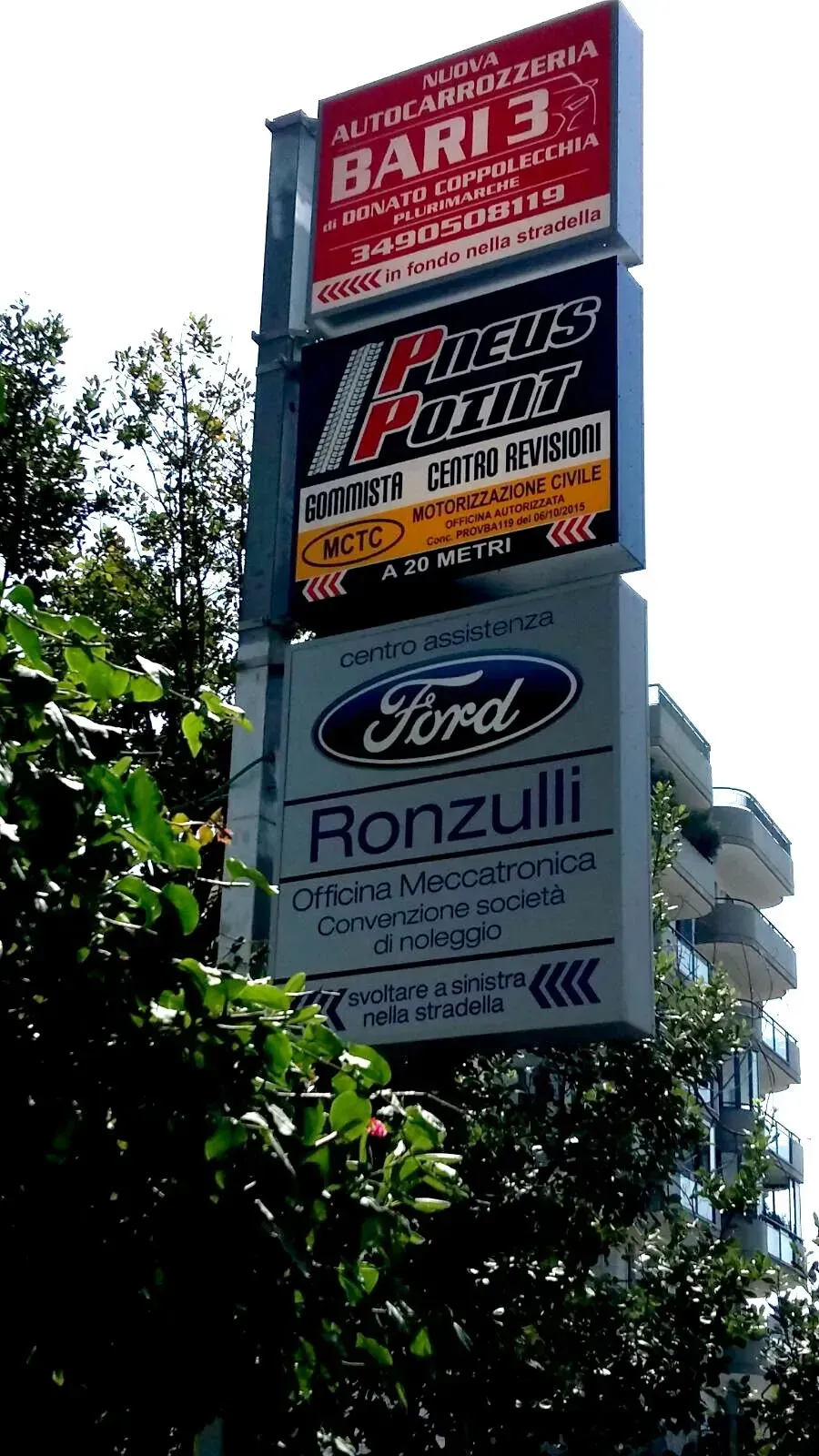 Alto cartellone pubblicitario a bordo strada con numerose inserzioni commerciali sullo sfondo di un cielo azzurro e alberi.
