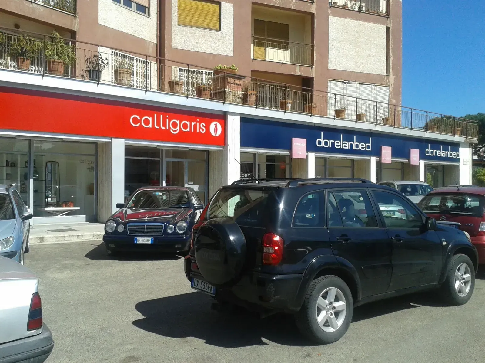 Vista stradale di auto parcheggiate davanti a negozi con cartelli rossi e blu sotto palazzi residenziali