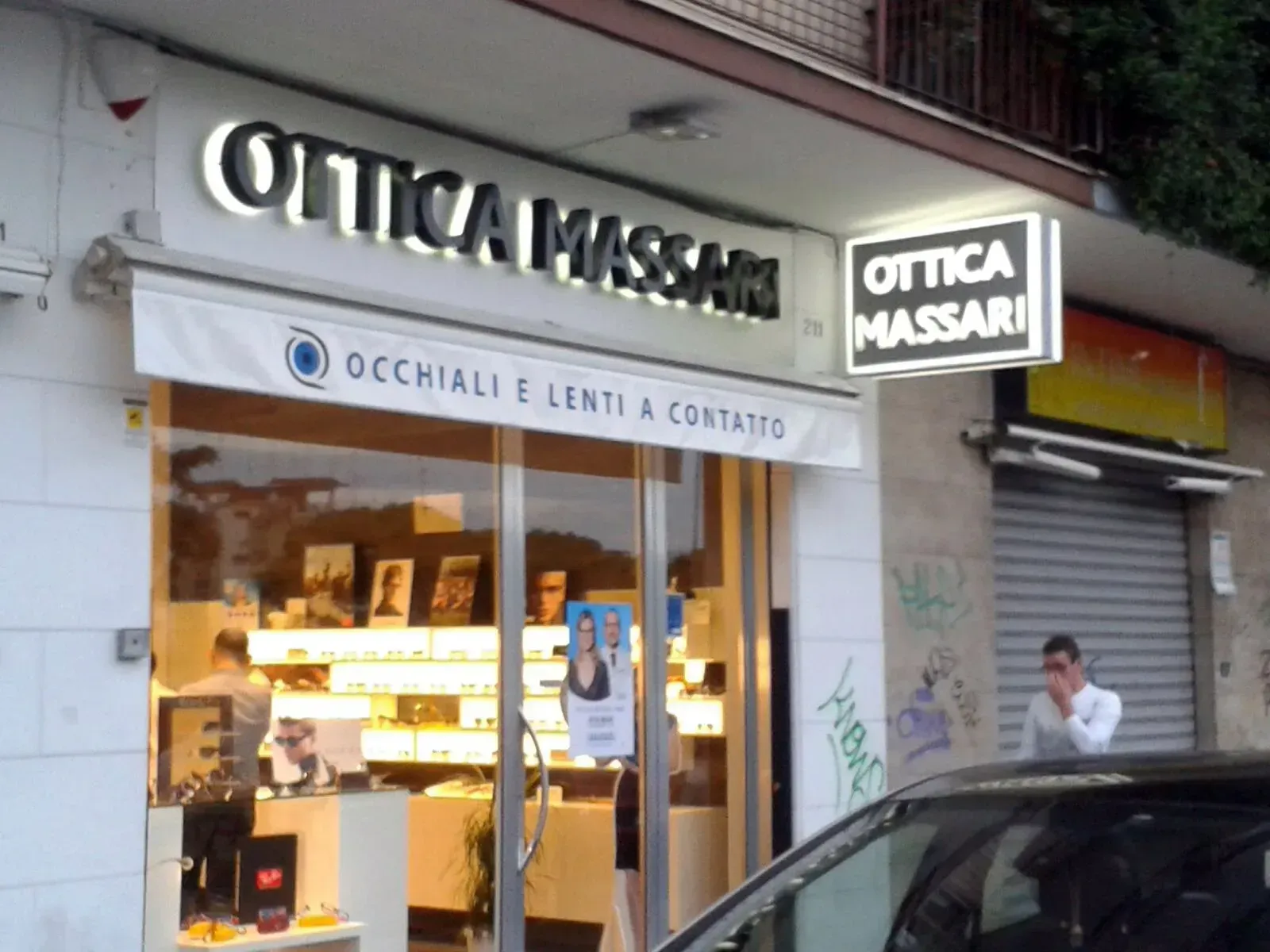 Vetrina del negozio di occhiali Ottica Massari con interni illuminati e una persona in piedi sul marciapiede all'esterno.