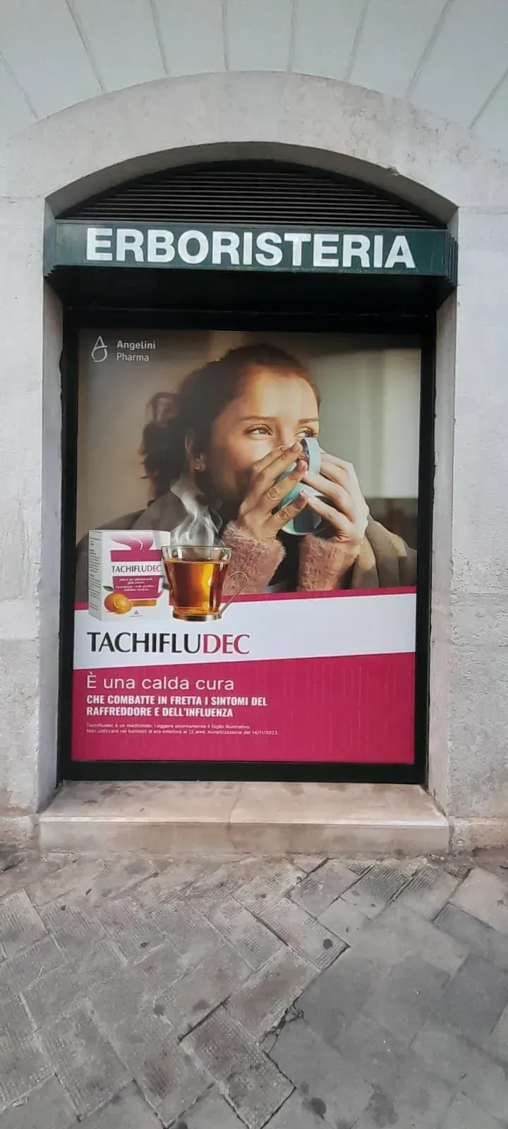 Vetrina del negozio Erboristeria con una grande pubblicità rosa di Tachiflux che mostra un bambino che beve da una tazza